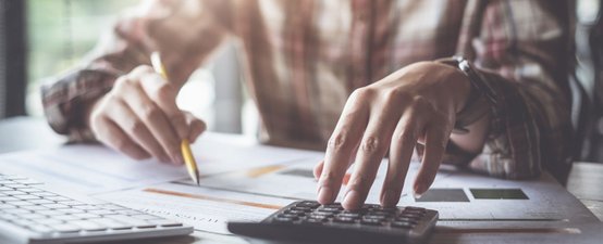 Eine Person mit einem karierten Hemd nutzt einen Taschenrechner, während sie auf Dokumente am Tisch schaut.