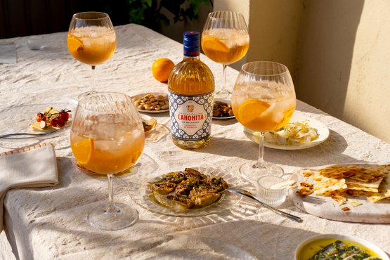 Ein Tisch gedeckt mit Gläsern voller Getränken, einer Flasche Canonita und verschiedenen köstlichen Speisen im Sonnenlicht.