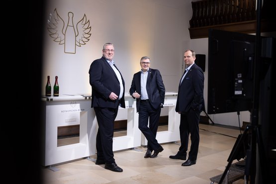 Drei Männer in Anzügen posieren lässig vor einem eleganten Hintergrund mit einem Logo und Flaschen Sekt auf einem Tisch.