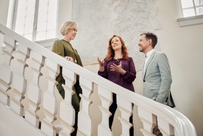 Three people are standing on a staircase, having a lively conversation while smiling at each other.