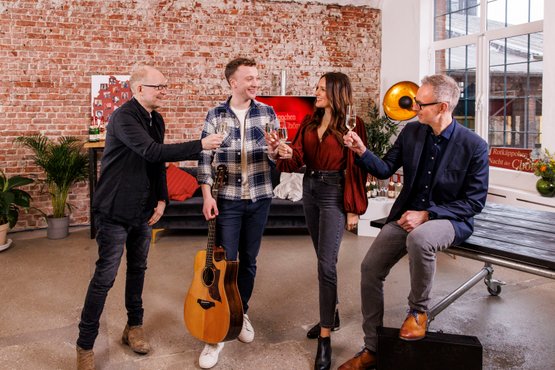 Vier Personen stoßen mit Gläsern an, während einer mit einer Gitarre in der Hand lächelt, in einem modernen Raum mit Backsteinwand.