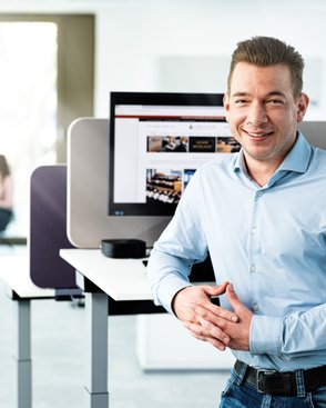 Ein lächelnder Mann in einem hellen Büro steht an einem Stehpult, während zwei Frauen im Hintergrund in einem Gespräch vertieft sind.