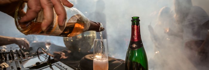 Ein DJ-Pult im Vordergrund, während ein Glas mit sprudelndem Sekt gefüllt wird, umgeben von verschwommenen Feiernden.