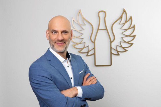Markus Jauch: The man with a gray beard stands smiling in a blue suit, while behind him the Rotkäppchen Mumm logo, an artistic wing motif, can be seen.
