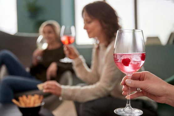 Eine Hand hält ein Glas mit pinkem Getränk, während zwei Frauen im Hintergrund anstoßen und entspannen.