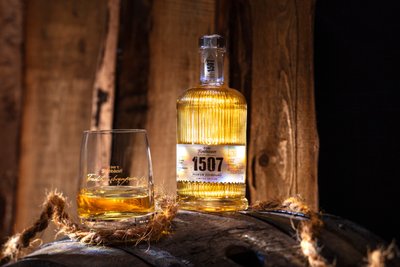 Die Flasche mit der Aufschrift '1507' und ein Glas mit goldenem Kornbrand sind auf einem Holzfass in einer gemütlichen Umgebung arrangiert.