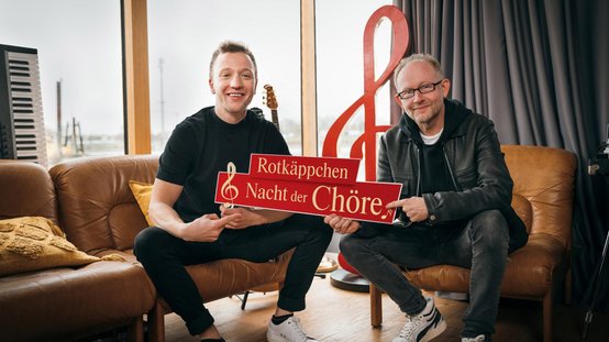 Zwei Männer sitzen auf einer Couch und halten ein rotes Schild mit der Aufschrift 'Rotkäppchen Nacht der Chöre'.