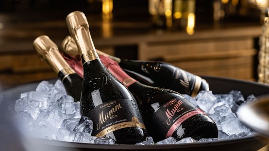 Drei Flaschen Mumm-Sekt liegen auf Eis in einer eleganten Schüssel, umgeben von glitzerndem Wasser und einer warmen Holzumgebung.