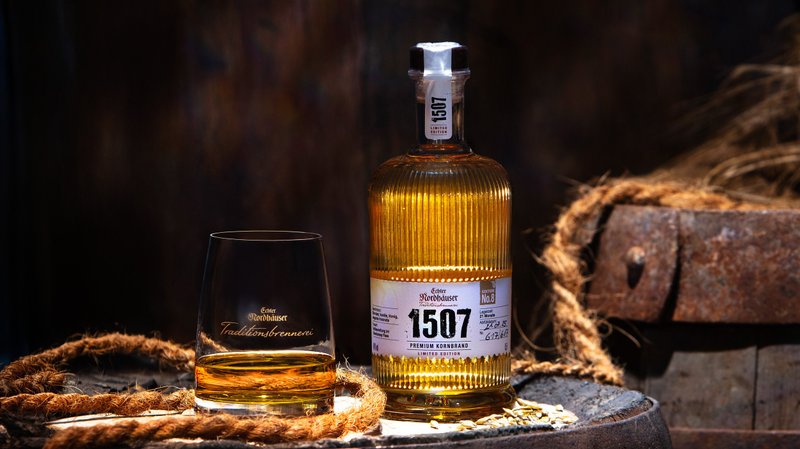 Eine Flache Echter Nordhäuser Traditionsbrennerei 1507 No. 8 und ein Glas stehen auf einem Holzfass vor dunklem, rustikalem Hintergrund.