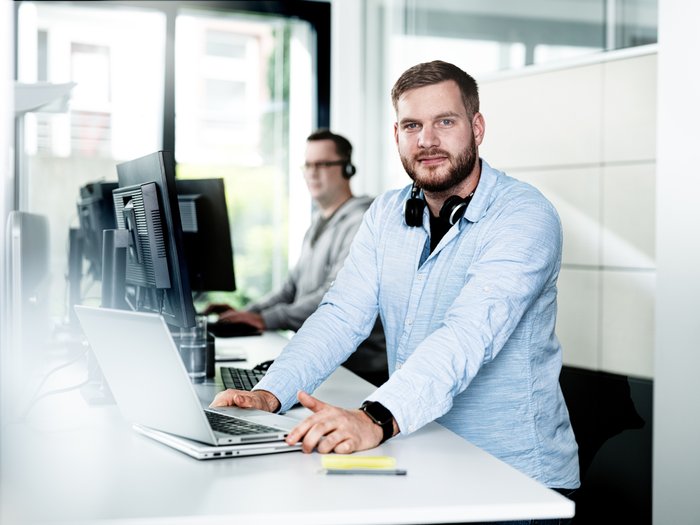 The man in the foreground is wearing a light blue shirt and is looking directly into the camera while working on his laptop.