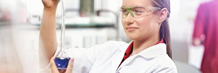 Eine junge Frau in einem Laborkittel hält eine Pipette mit einer blauen Flüssigkeit und trägt Schutzbrille.