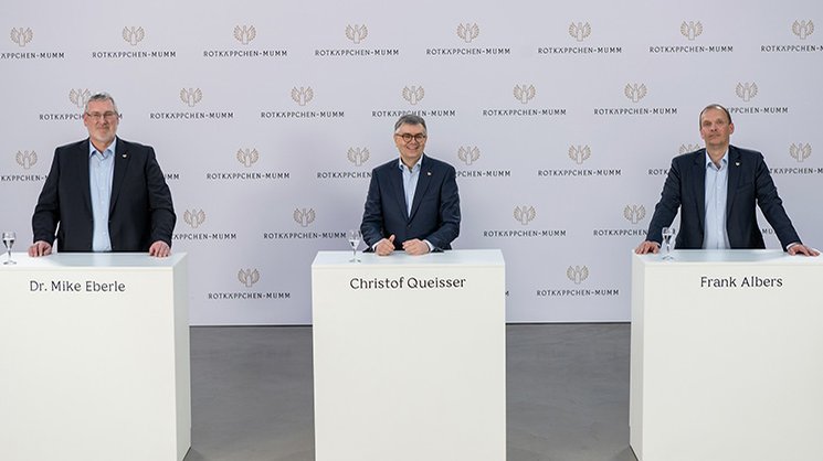 Drei Männer stehen an weißen Tischen, lächeln und präsentieren sich vor einer neutralen Wand mit dem Logo von Rotkäppchen-Mumm.