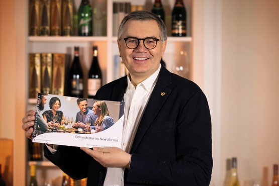 Ein lächelnder Mann hält ein Buch mit dem Titel 'Genusskultur im New Normal' vor einer Bar mit Weinflaschen im Hintergrund.