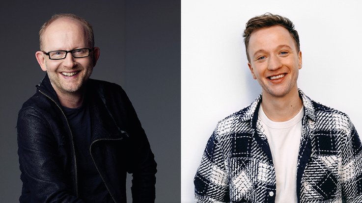 Zwei Männer stehen nebeneinander, einer mit Brille und lächelnd, der andere in einem karierten Hemd, beide wirken freundlich.