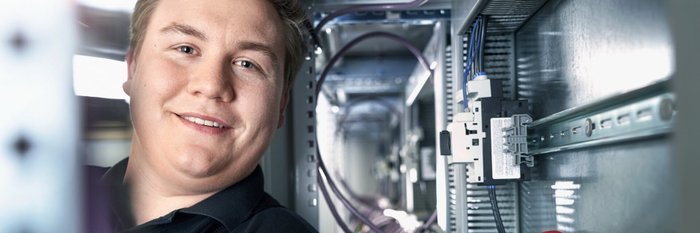 Ein junger Mann lächelt zufrieden, während er in einem technischen Raum mit Kabeln und Geräten arbeitet.