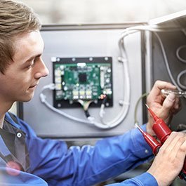 Ein junger Techniker arbeitet konzentriert an einem elektrischen Gerät, während er mit Messgeräten Kabel überprüft.