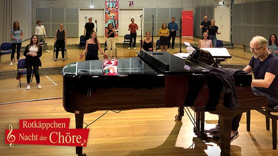 Ein Pianist sitzt am Klavier, während eine Gruppe von Sängern in verschiedenen Posen für eine Aufführung probt.