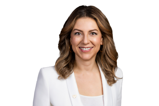 Silvia Wiesner: A smiling woman with long, curly hair wears a white blazer and gold earrings against a dark background.