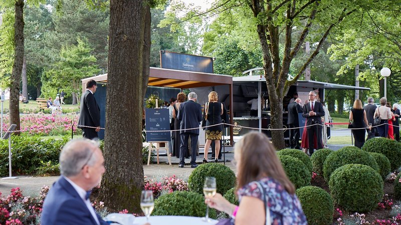 Elegante Gäste unterhalten sich in einem Park, während sie an einem Getränkestand stehen, umgeben von bunten Blumenbeeten.