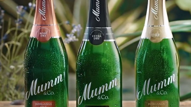 Drei Flaschen Mumm Sekt stehen auf einem Holztisch, umgeben von grünen Pflanzen und lila Blumen im Hintergrund.