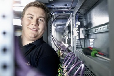 A young technician smiles contentedly while working in a control cabinet with colorful cables.