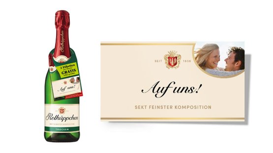 Das Etikett der Sektflasche zeigt den Schriftzug 'Auf uns!' und ein Bild eines glücklichen Paares, das sich anlächelt.