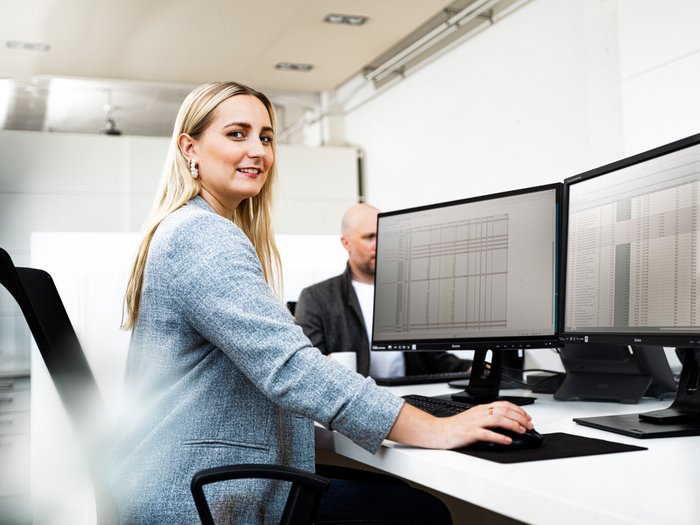 A blonde woman sits at a desk with two monitors, smiling and working concentratedly on a computer.