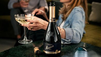Eine elegante Flasche Godefroy Mumm Champagner steht auf einem Tisch, während eine Person ein Glas Sekt hält und anstößt.