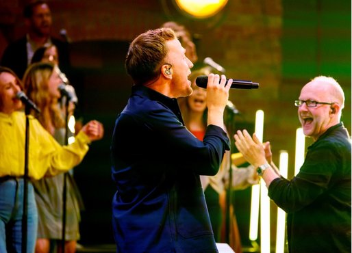 Ein Sänger mit Mikrofon performt leidenschaftlich auf der Bühne, während die Band im Hintergrund begeistert mitwirkt.