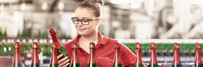 The employee in a red shirt inspects a champagne bottle while standing in a factory full of green bottles.