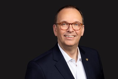 Frank Albers: A smiling man with glasses wears a dark suit and a white shirt, against a neutral background.
