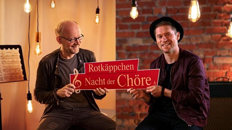Zwei Männer sitzen in einem gemütlichen Raum, lächelnd und halten ein Schild mit der Aufschrift 'Rotkäppchen Nacht der Chöre'.