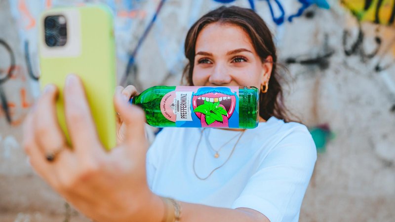 Mit einem fröhlichen Ausdruck hält die Frau eine Flasche Pfeffi in der Hand, während sie ein Selfie vor einer bunten Wand macht.