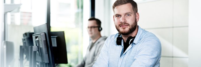 Der Mann im Vordergrund trägt ein hellblaues Hemd und schaut direkt in die Kamera, während er an seinem Laptop arbeitet.