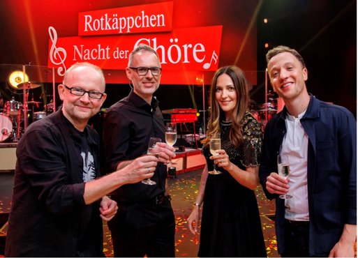 Vier Personen stehen lächelnd zusammen und stoßen mit Sektgläsern an, während bunte Konfetti auf dem Boden liegt.