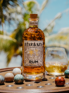 Eine Flasche Star & Key Rum steht auf einem Tisch neben einem Glas mit Eiswürfeln, im Hintergrund sind Palmen zu sehen.