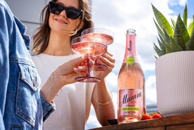 Zwei Frauen stoßen mit gläsernen Sektgläsern an, während eine Flasche alkoholfreier Rosé im Vordergrund steht.