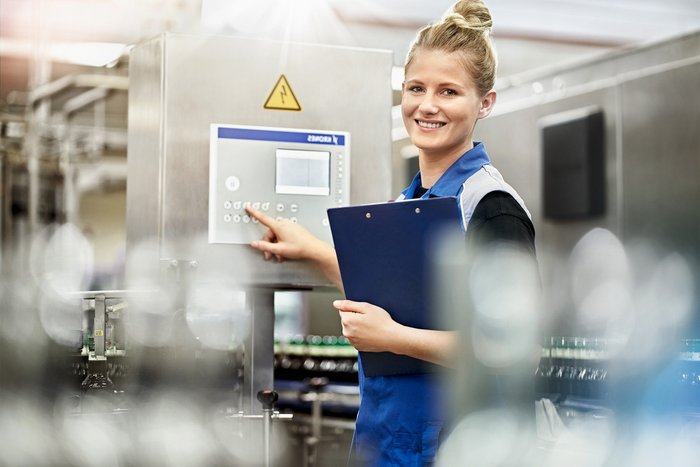 Eine lächelnde Frau in Arbeitskleidung bedient ein Bedienfeld in einer modernen Fabrikumgebung.