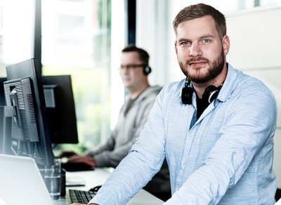 Ein junger Mann mit Bart und Kopfhörern lächelt in einem modernen Büro, während ein Kollege im Hintergrund arbeitet.