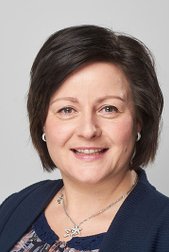 Eine lächelnde Frau mit dunklen, schulterlangen Haaren trägt eine elegante Bluse und eine Kette mit einem Sternanhänger.