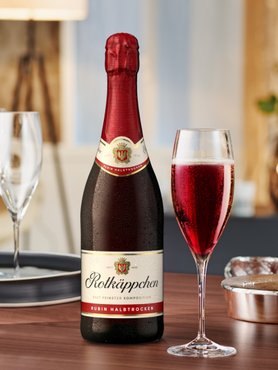 Eine Flasche Rotkäppchen Sekt steht auf einem Holztisch, daneben ein Glas mit sprudelndem, rubinrotem Getränk.