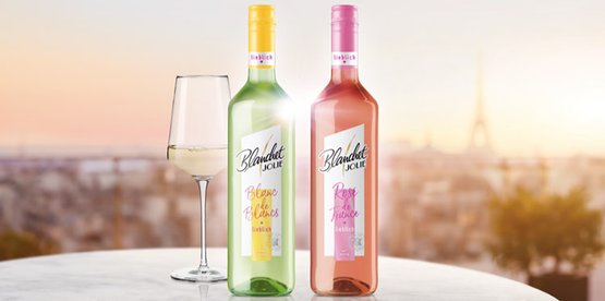 Zwei Flaschen Wein Blanchet stehen auf einem Tisch, eine grüne und eine rosafarbene, mit einem Glas Weißwein daneben.
