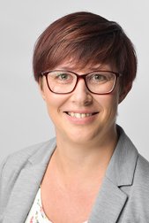 A smiling woman with short red hair and glasses wears a grey blazer and stands in front of a light background.