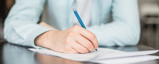 Ein Mann in einem hellblauen Hemd hält einen Stift und notiert etwas auf einem Blatt Papier.