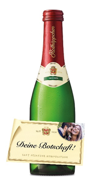 Eine Flasche Sekt mit rotem Kapselverschluss steht im Vordergrund, begleitet von einer festlichen Botschaft auf einer Karte.