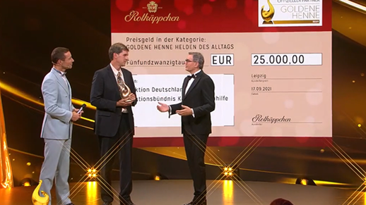 Drei Männer stehen auf einer Bühne, während einer einen Preis für die Goldene Henne entgegennimmt, umgeben von festlicher Beleuchtung.