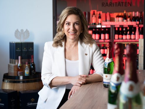 Silvia Wiesner an einem Holztisch mit Sektflaschen, im Hintergrund Weinregale mit Raritäten.