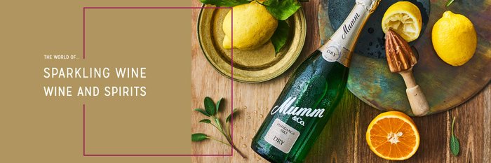A bottle of Mumm sparkling wine stands next to fresh lemons and oranges on a rustic wooden base.