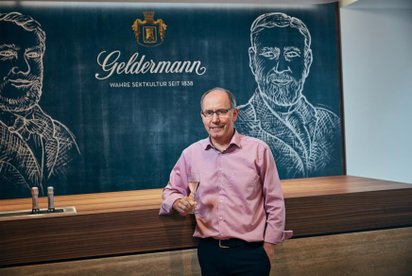 Ein Mann in einem rosa Hemd hält ein Glas Sekt und lächelt vor einer Wand mit einer Zeichnung und dem Schriftzug 'Geldermann'.