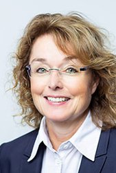 A smiling woman with curly brown hair wears glasses and a dark blazer against a light background.